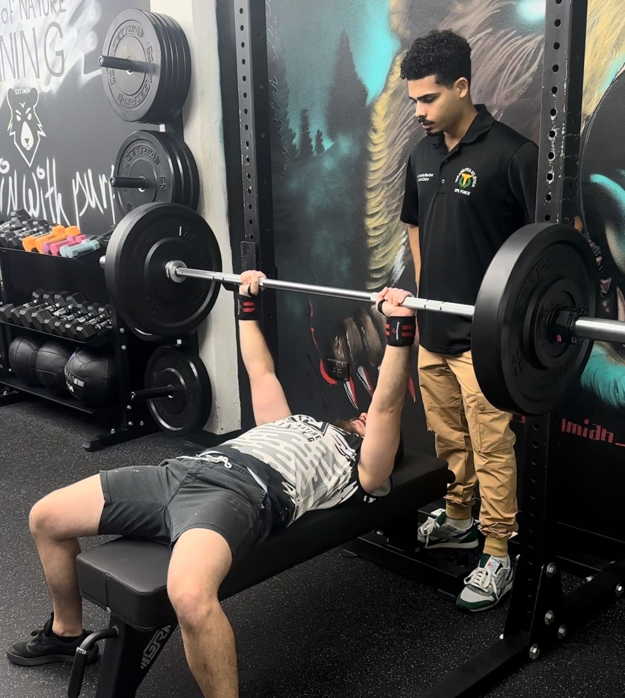 Man bench pressing in gym with a spotter standing nearby, surrounded by weights and gym equipment.