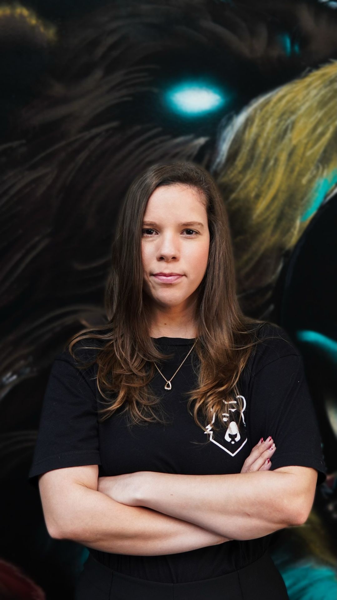 Person wearing a black t-shirt with crossed arms standing in front of a dark, glowing mural.